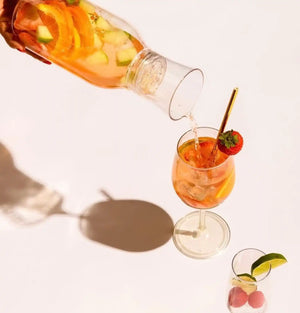 Clear glass pitcher pouring fruit-infused drink into a stemmed glass.